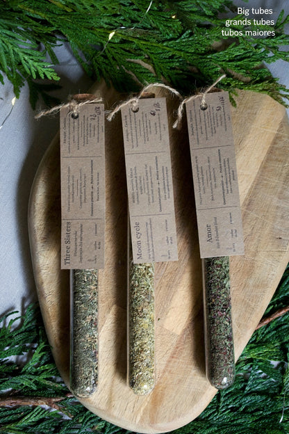 3 glass tubes containing teas and infusions, with their kraft label on top. On a wooden board and surrounded by greenery