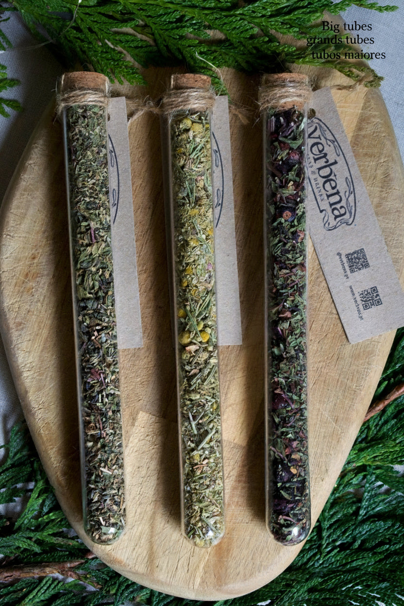 3 glass tubes containing teas and infusions, with their kraft label below them. On a wooden board and surrounded by greenery