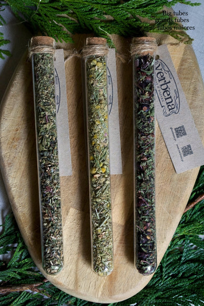 3 glass tubes containing teas and infusions, with their kraft label below them. On a wooden board and surrounded by greenery