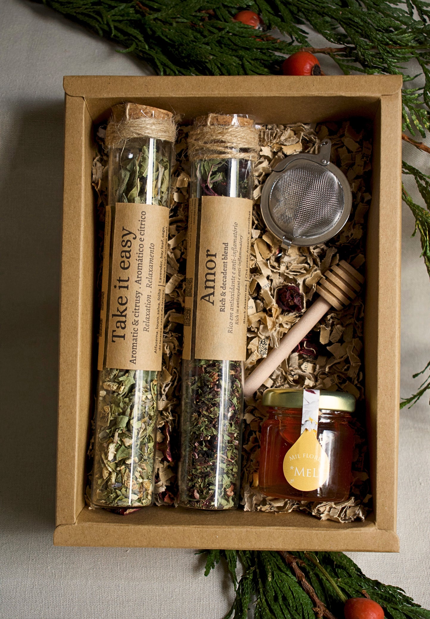 Herbal tea set in glass tubes, a strainer, and honey in a cardboard box with greenery in the background.