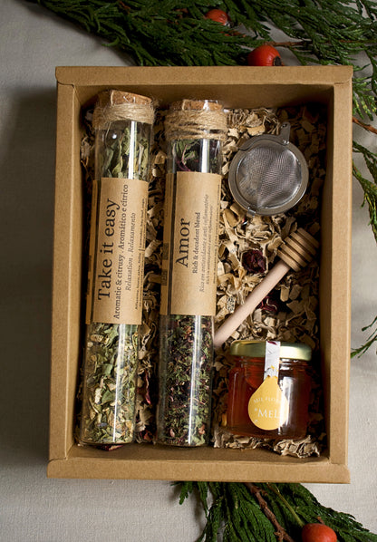 Herbal tea set in glass tubes, a strainer, and honey in a cardboard box with greenery in the background.