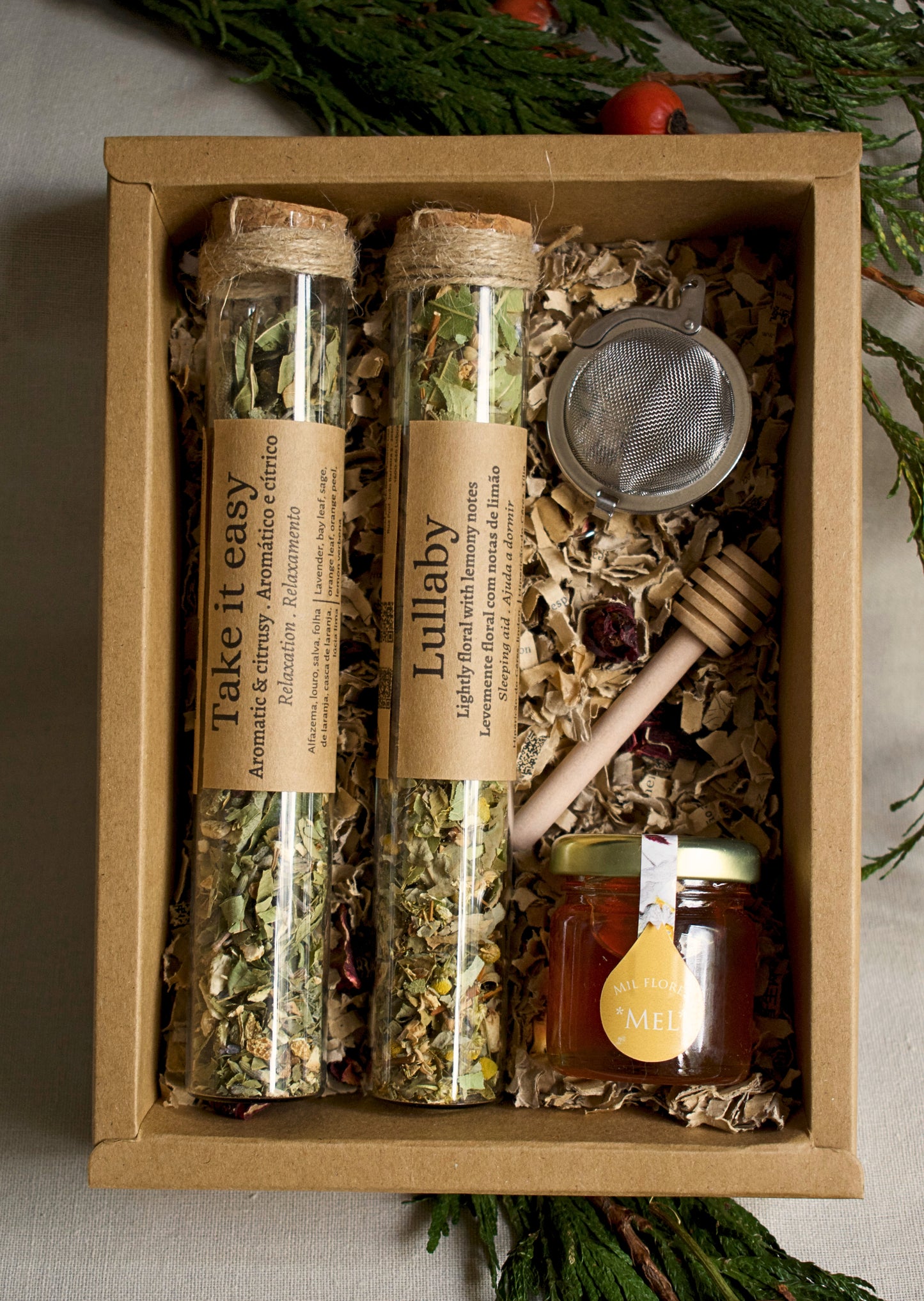 Herbal tea set in glass tubes, a strainer, and honey in a cardboard box with greenery in the background.
