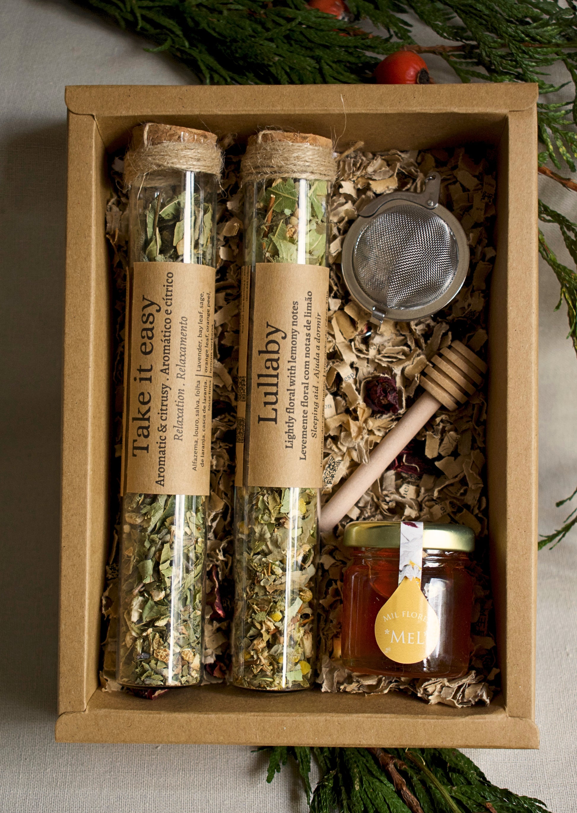 Herbal tea set in glass tubes, a strainer, and honey in a cardboard box with greenery in the background.