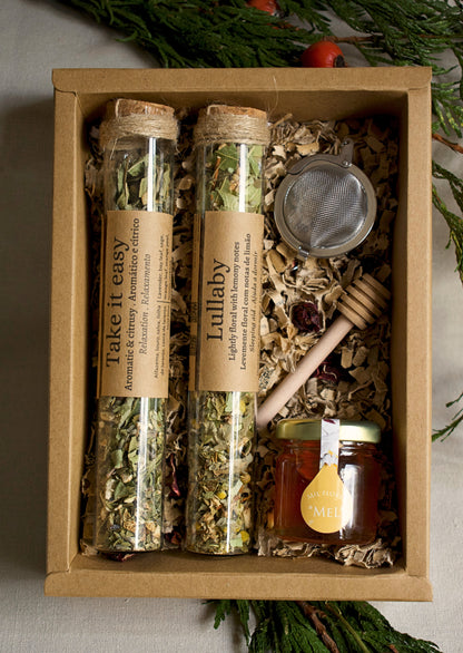 Herbal tea set in glass tubes, a strainer, and honey in a cardboard box with greenery in the background.