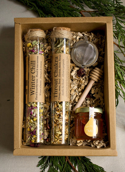 Herbal tea set in glass tubes, a strainer, and honey in a cardboard box with greenery in the background.