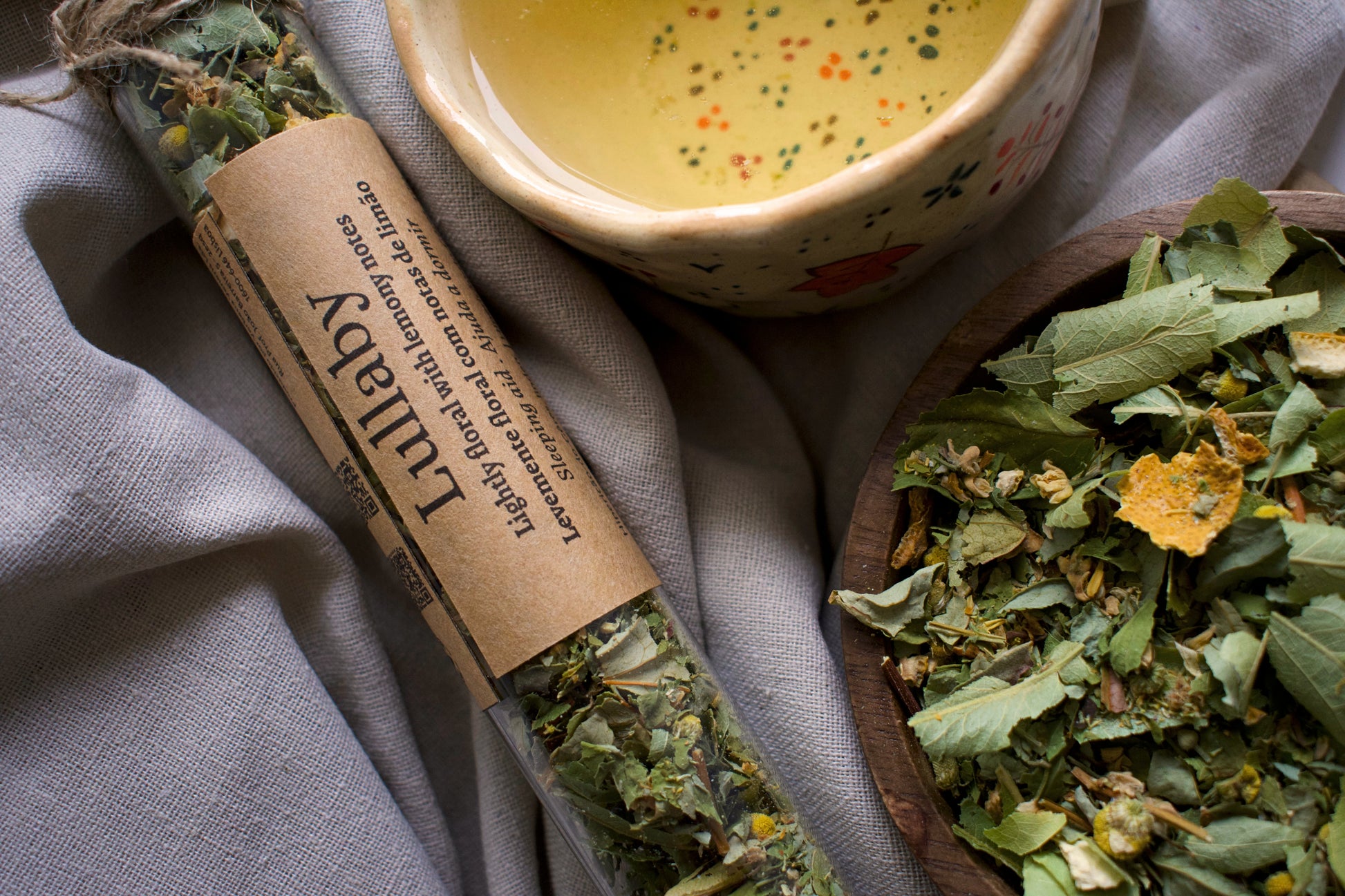 In situ, glass tube with herbal blend lullaby with a cup of tea and wooden bowl filled with the herbal blend on a linen cloth. 