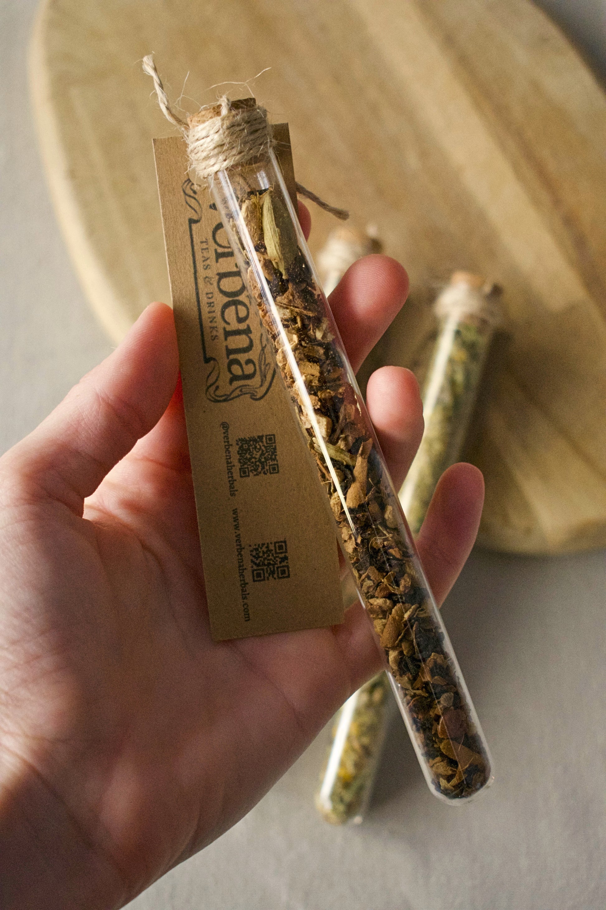 Glass tube held by a hand, filled with tea blend and has a kraft label attached to it. Background: wooden board and linen tablecloth
